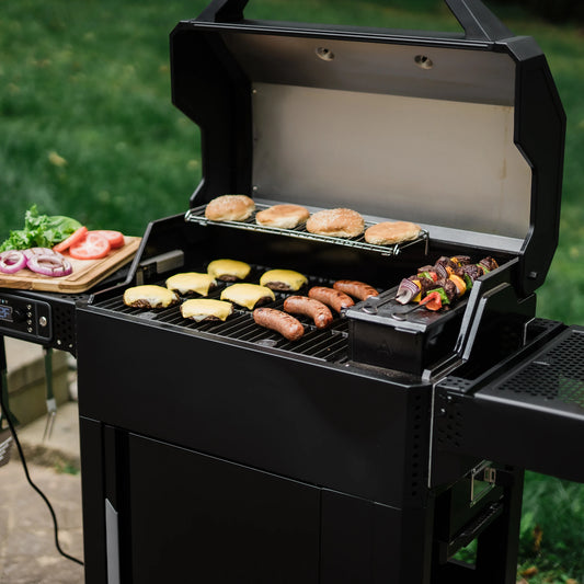 An open AutoIgnite grill with cheeseburgers and sausages cooking on the grill, kebabs searing on the charcoal hopper sear plate, and buns toasting on the warming rack
