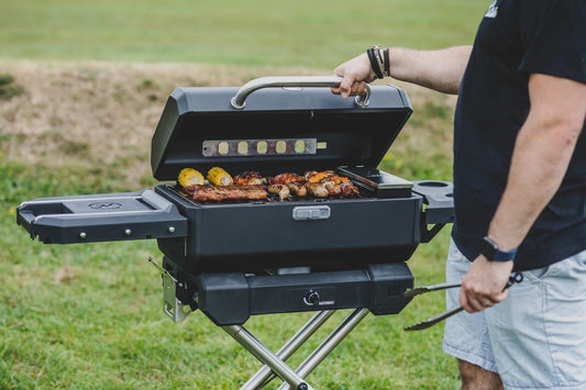 A man wearing a black t-shirt and pale blue shorts opens a Masterbuilt portable charcoal grill on a QuickConnect cart. Sausages, kabob skewers, chicken breasts, steaks, and corn on the cob cook on the grill.