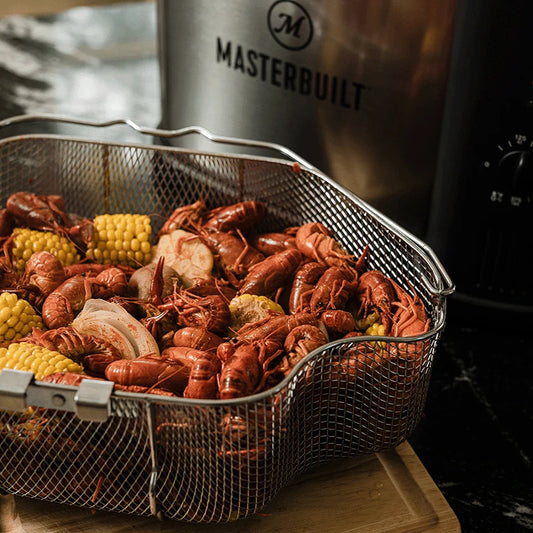 A steamed low country boil with shrimp, corn and crawfish sits draining in a fryer basket placed on a grooved board next to a Masterbuilt 10L fryer
