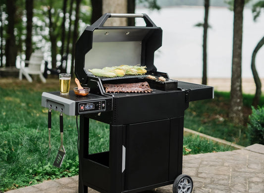 An open AutoIgnite 545 sits on a paved patio near a lake. Husk-on corn on the cob roasts on the warming rack while racks of ribs grill on the main cooking grate.