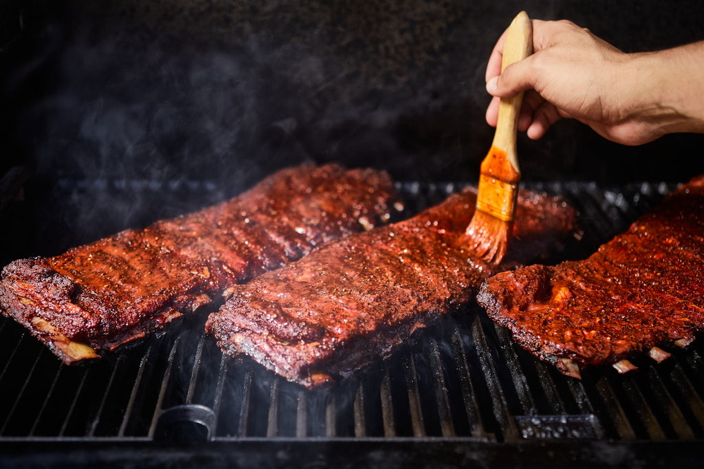 A person uses a wood-handled brush to spread barbecue sauce on 3 racks of ribs smoking on grill grates