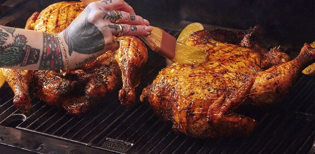 A man with tattoos on hand and arm uses a brush to base turkeys roasting on a grill grate