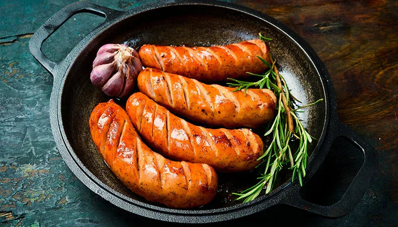 4 plump and juicy smoked sausages, all with deep cut marks in them, sit in a cast iron pan with a sprig of rosemary and a whole garlic bulb.