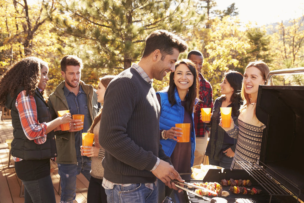 A diverse crowd of young adults gather near a grill during autumn. Most of the people are wearing cool-weather clothing and holding drink cups. One man stands in the foreground cooking kebabs and burgers in an open grill.