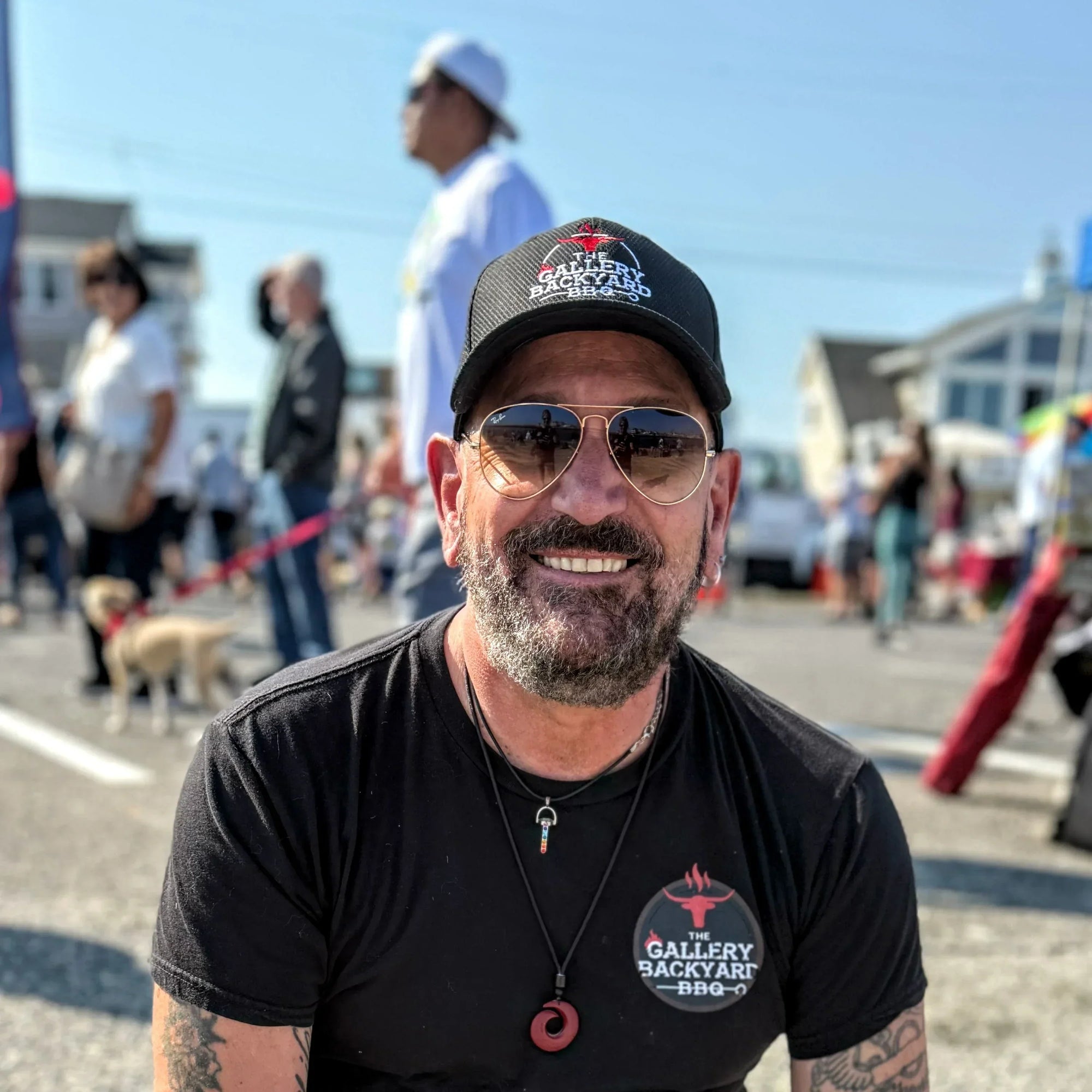 Ambassador Thomas Scarano outside on a sunny day, surrounded by  people. He is wearing a black t-shirt and black hat, both with the Gallery Backyard BBQ logo in red and white 