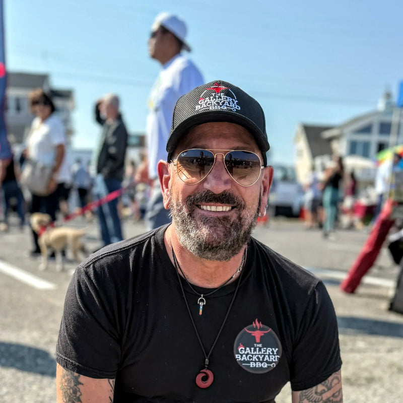 Ambassador Thomas Scarano outside on a sunny day, surrounded by  people. He is wearing a black t-shirt and black hat, both with the Gallery Backyard BBQ logo in red and white 