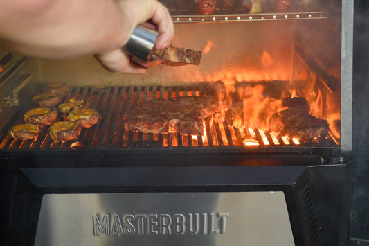 A hand holding tongs reaches into flames coming up through the cooking grate of a Gravity Series grill to flip steaks over. Several stuffed mushrooms sit on the grill grate to the left of the steaks. There is no direct fire under the mushrooms.