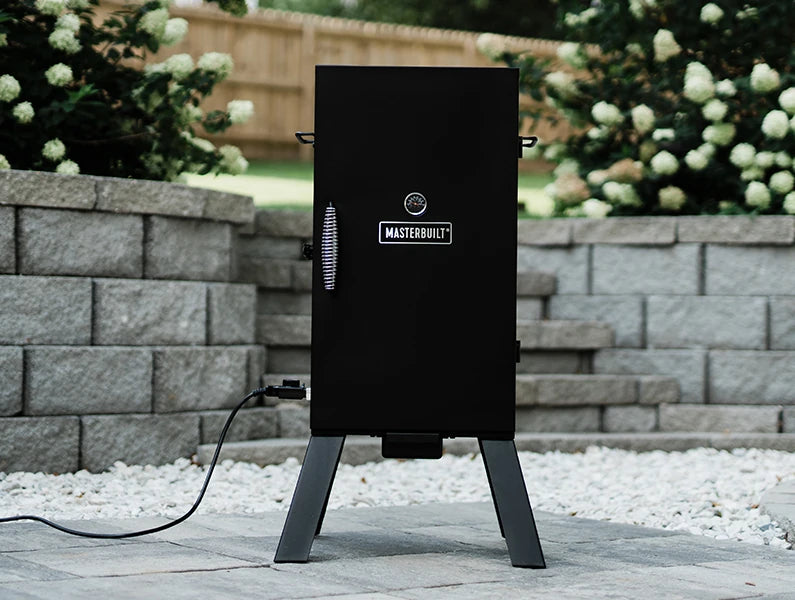 An analog electric smoker sits on a paved patio in front of stone stairs and a retaining-wall terrace. The smoker has a cool-touch wire handle, temperature gauge, and Masterbuilt name plate on the door. The smoker sits on legs. An electric cord with temperature control knob is plugged into the left side of the smoker.