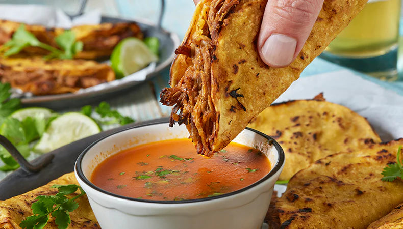 Hand dipping a taco into a bowl of red sauce with other tacos and limes in the background.