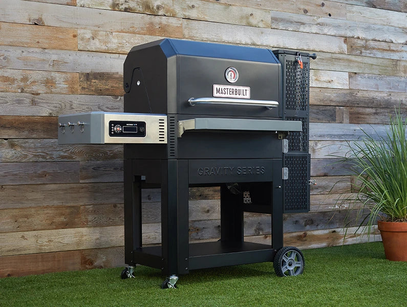 A Gravity Series grill sits in front of a wooden wall. The grill has a shelf with control panel on the left, charcoal hopper on the right, and sits on a wheeled cart.