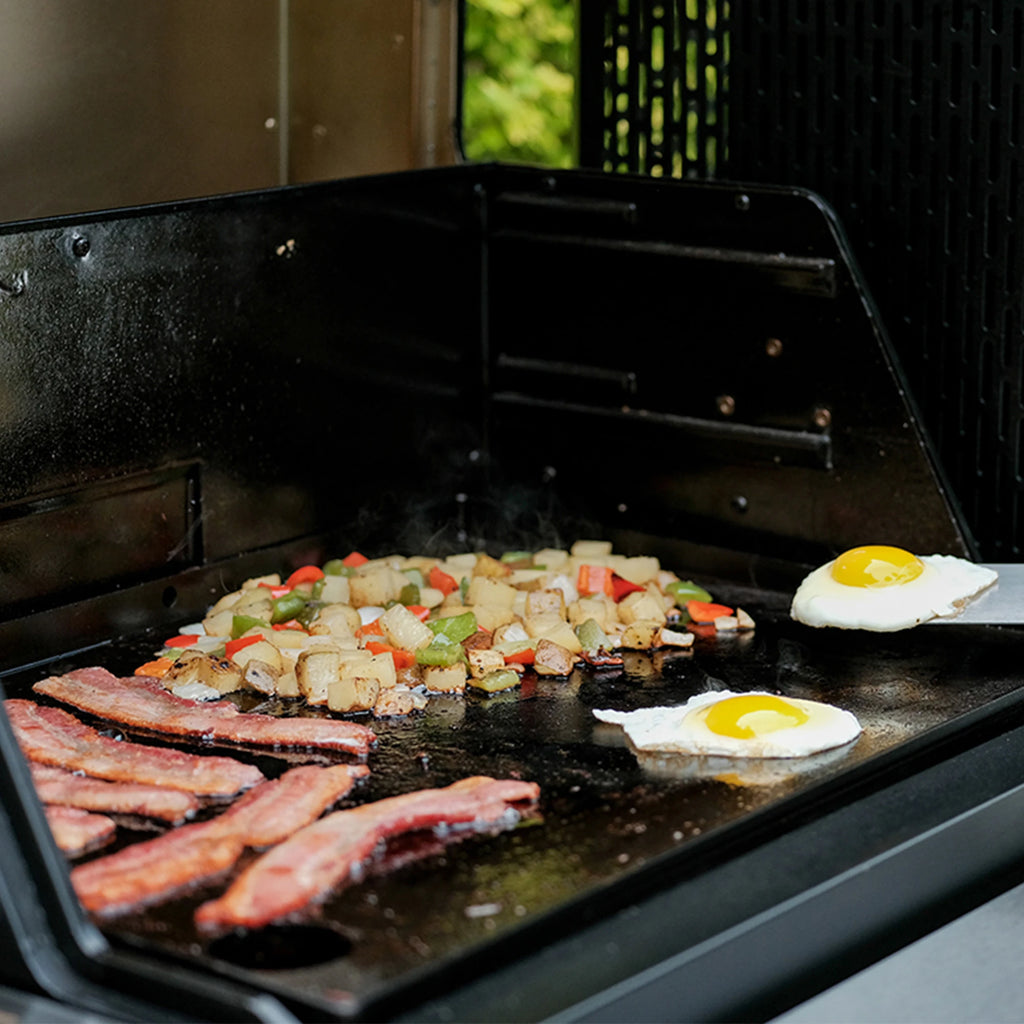 Grill with bacon, hashbrowns, and eggs cooking on a black surface.