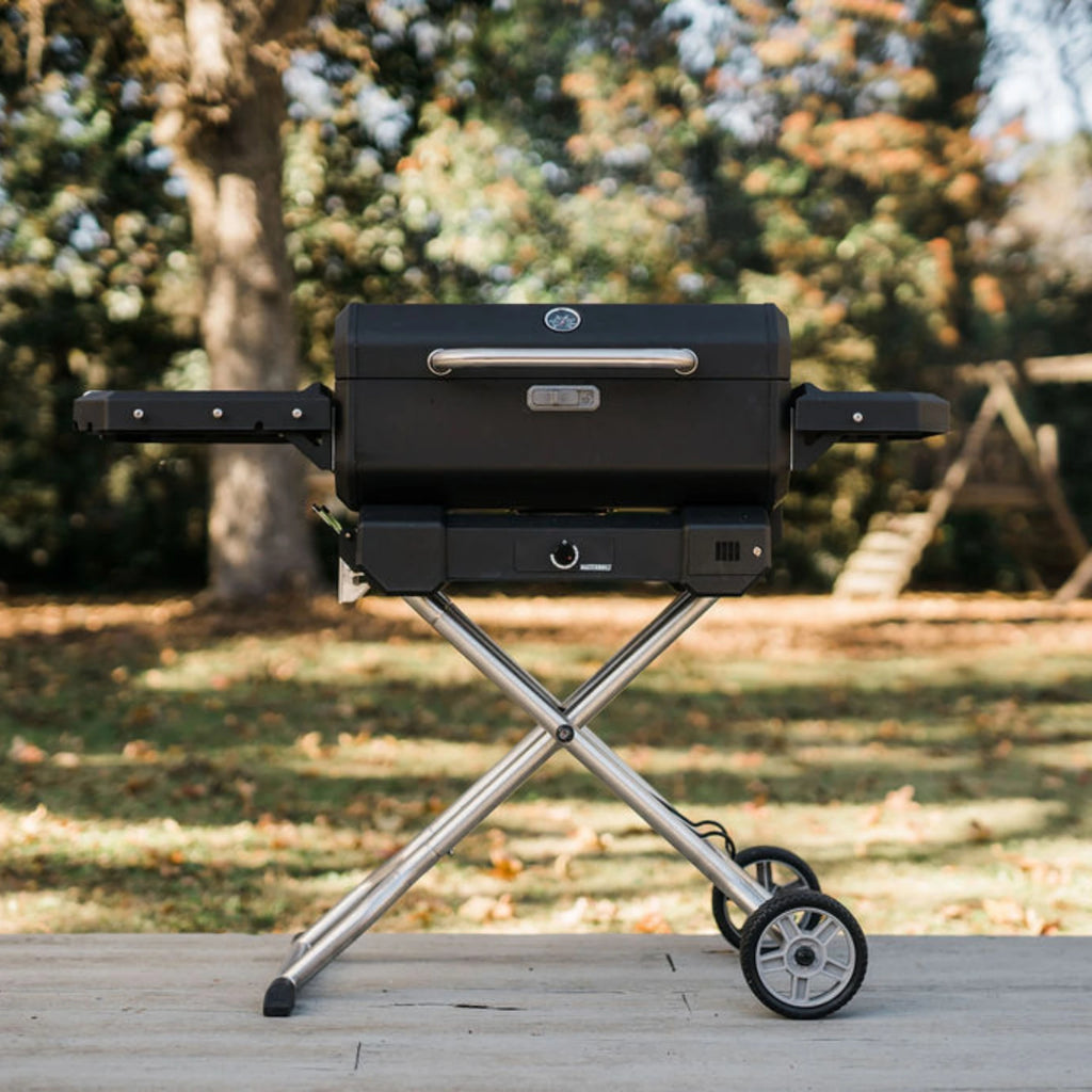 Masterbuilt Portable Grill with cart on a backyard deck. Trees and a wooden playset are visible in the background.