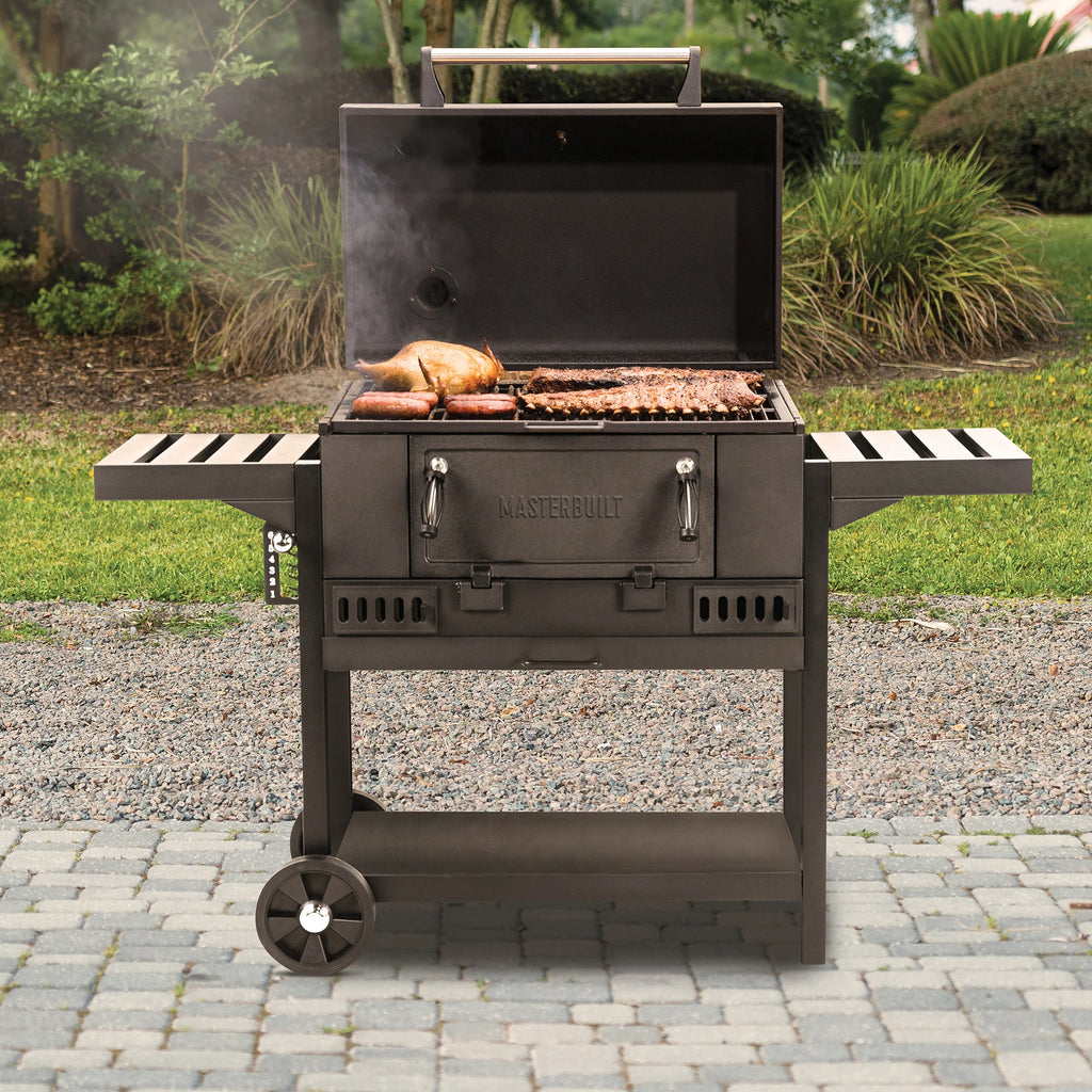 30" charcoal grill cooking ribs, chicken and hot dogs on a cobblestone patio in front of a gravel bed, lawn, and tropical bushes