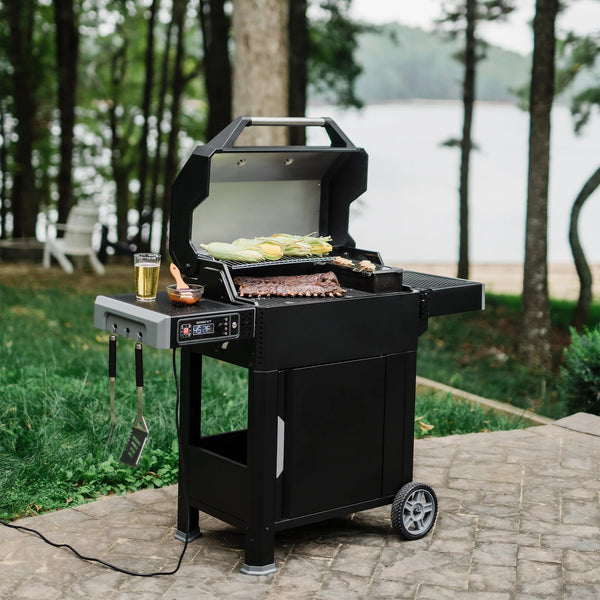 An open AutoIgnite grill on a patio in front of a wooded area leading to a beach and lakeshore. A rack of ribs cooks on the main grate while corn on the cob roasts on the warming rack and shrimp skewers sear on the hopper grill plate. Utensils hang from the left side shelf. A full drink glass plus a glass bowl of sauce with a brush resting in it sit on the side shelf.