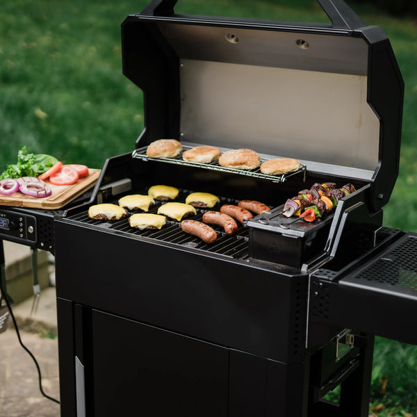 An open AutoIgnite Grill and Smoker. Cheeseburgers and sausages grill on the main cooking grate. 2 hamburger buns toast on the warming rack, and 2 skewers rest on the QuickSear hopper cast iron griddle plate. A wooden cutting board with lettuce leaves, sliced tomatoes, and grilled sliced onions is on the left side shelf. 