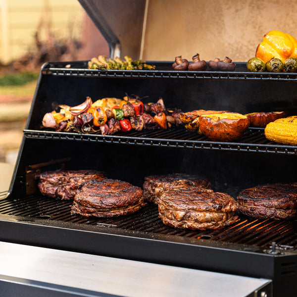 Open Gravity Series 1050 with steaks searing on main cooking grate. Lower warming rack holds kebabs, acorn squash halves, and corn on the cob. Top warming rack has asparagus spears, whole mushrooms, Brussel sprouts, and a whole yellow bell pepper.