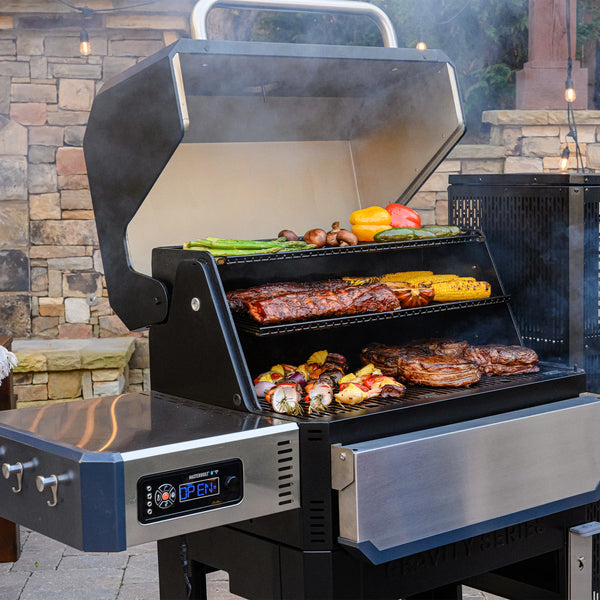 Open Gravity Series 1050 on an outdoor patio. A variety of food from steaks and kebabs to fresh vegetables fill the grill. The folding front shelf is in the down position. The left side shelf has built-in utensil racks on the outer edge.