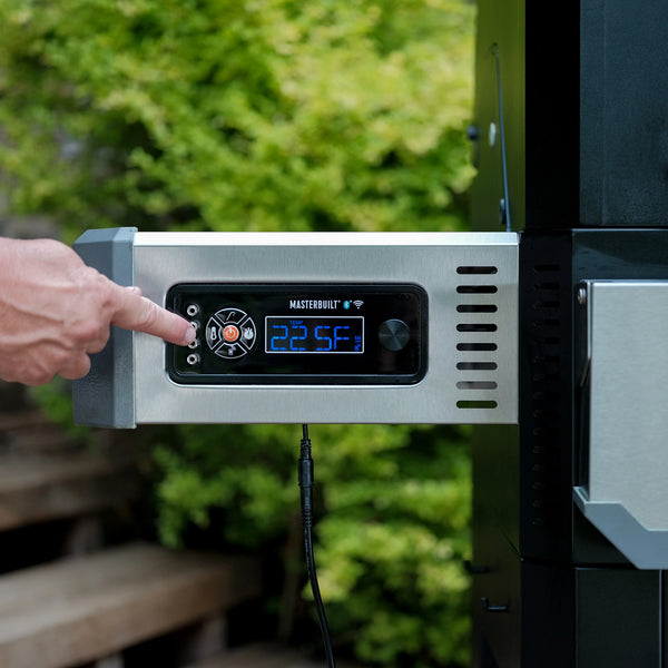 Digital control panel on a Gravity Series grill with a hand adjusting settings, set against a blurred green outdoor background. 
