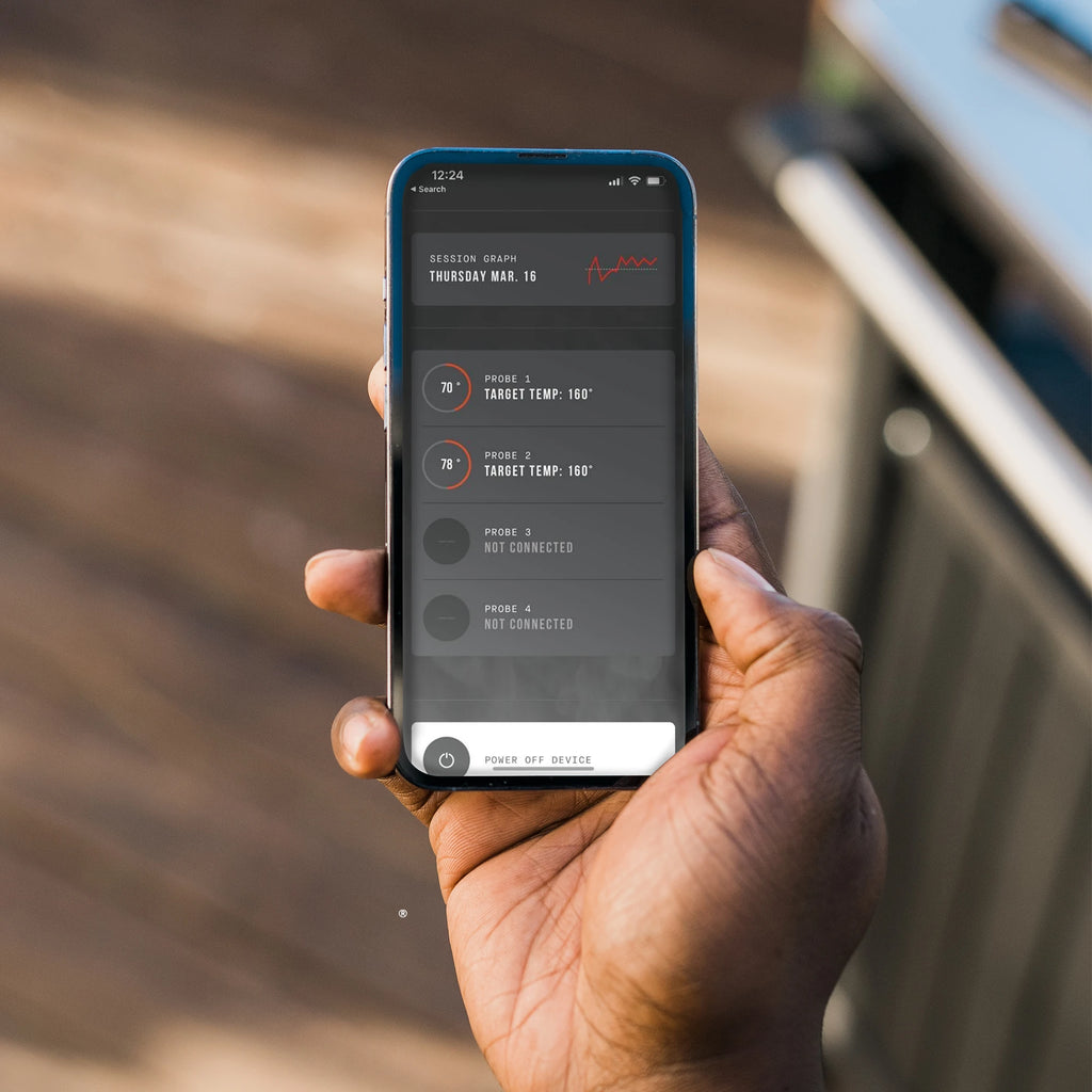 Hand holding a smartphone with Masterbuilt app control displayed, set against a blurred outdoor background. 