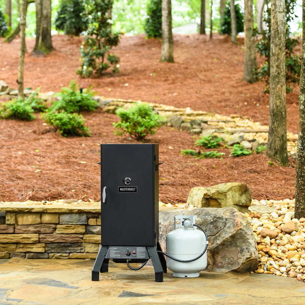 Black propane vertical smoker on a stone patio with a propane tank in a garden setting