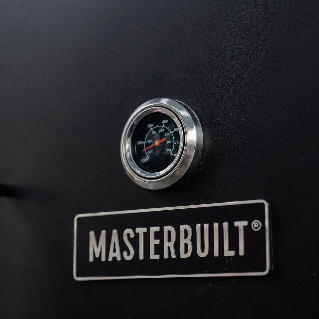 Closeup of the round temperature gauge in the top door with a Masterbuilt nameplate below. The gauge has both Fahrenheit and Celsius.