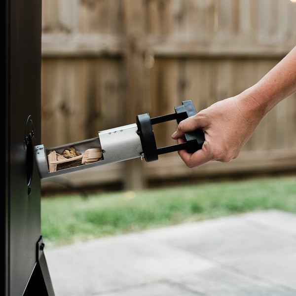 A man uses the side woodchip loader to add wood chips to the smoker. The loader is a horizontal metal cylinder with an opening to add the chips. The cylinder has a black plastic handle used to push, pull, and turn the loader.