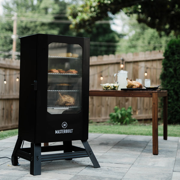 Black smoker with legs and a window in the door sitting on an outdoor patio in front of a table with food and drinks on it. Salmon, corn on the cob, macaroni and cheese and a whole chicken are visible through the smoker window.