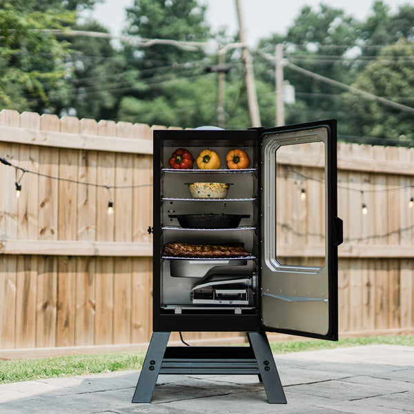 A smoker full of food sits on a patio in front of a wooden privacy fence strung with retro-style lights. The smoker is open to show 4 racks of food above the water and wood chip trays.