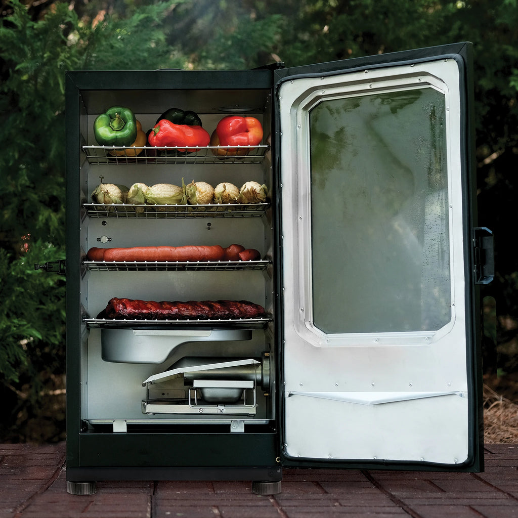 A Masterbuilt electric smoker with its door open, showing shelves with peppers, corn, sausage, ribs, and a removable water tray inside, set on a brick patio with trees in the background.