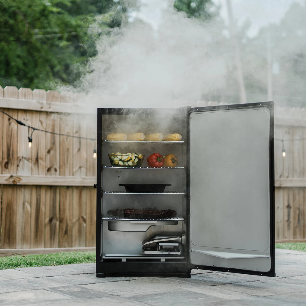 A smoker full of food sits on a patio in front of a wooden privacy fence strung with retro-style lights. The smoker is open to show 4 racks of food above the water and wood chip trays.
