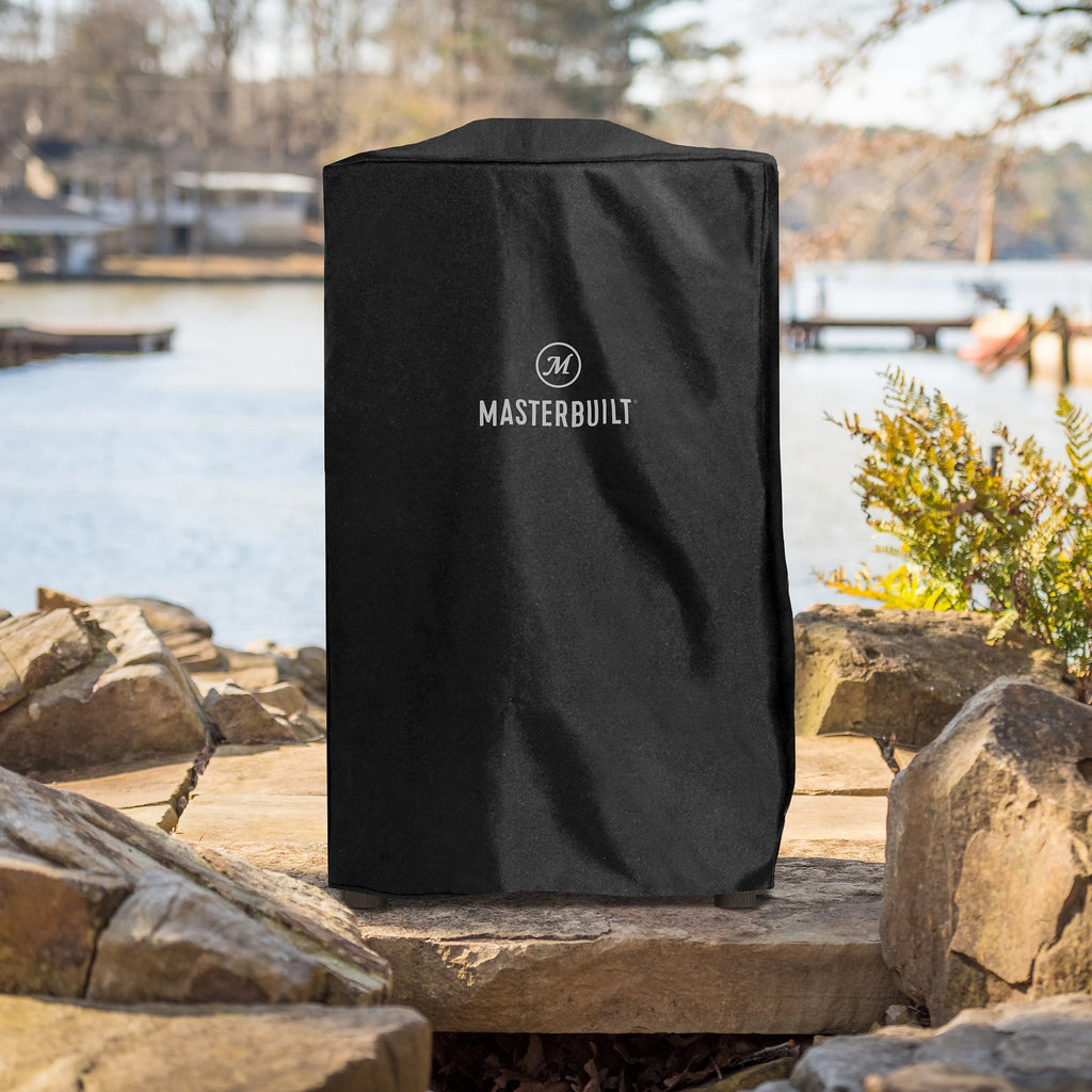 A black Masterbuilt smoker with cover stands outdoors on a stone surface with a lake and dock in the background.