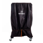 Black cover with double-stitched orange seams, an orange cinch cord at the bottom, and the Masterbuilt name and logo printed in white near the top. The grill is installed over a vertical smoker. Only the wheels on the bottom of the smoker cart are visible.