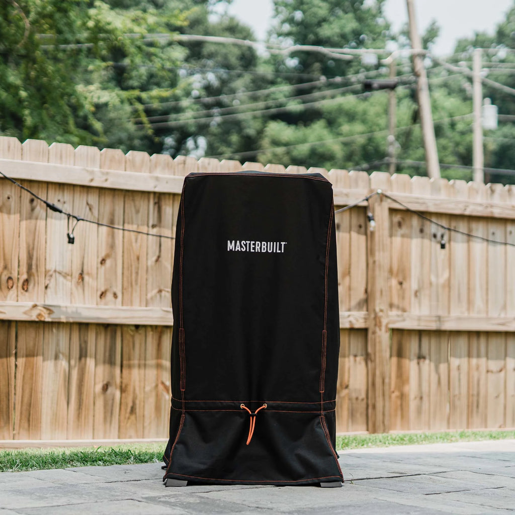 The cover installed over a vertical smoker with legs that is sitting outside on a paved patio in front a wooden privacy fence strung with lights.