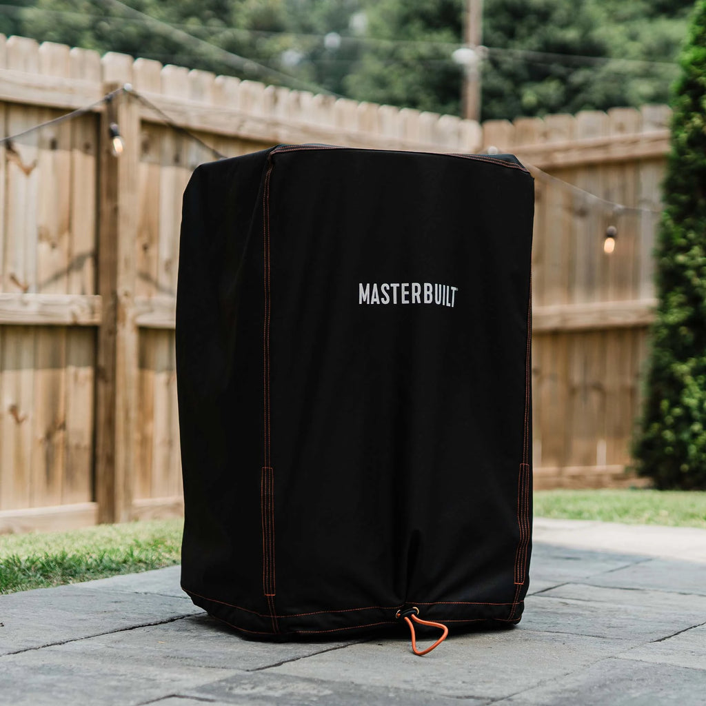 A covered vertical smoker without legs sits on a paved patio in front of a lawn and a wooden privacy fence strung with lights.