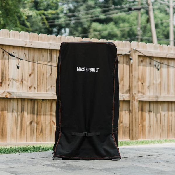 Vertical smoker on an outdoor patio in front of a wood privacy fence strung with decorative lights. The smoker is covered with a small universal smoker cover. The Masterbuilt® name mark is on the front of the cover. The seams at the corners are double-sticked with orange thread. 