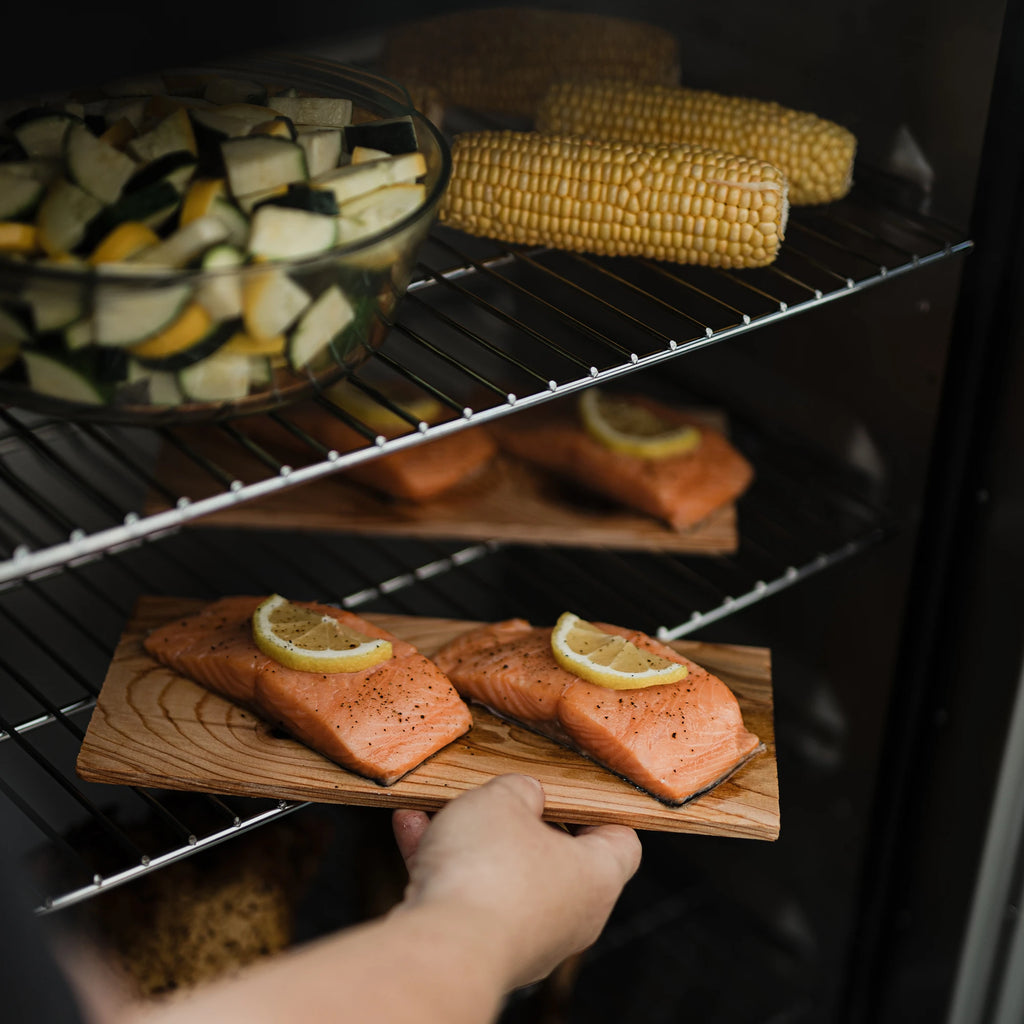 Some places salmon filets topped with lemon slices and placed on wood boards into a smoker. Already in the smoker are more salmon filets, corn on the cob, and a glass bowl of sliced yellow squash and zucchini. 