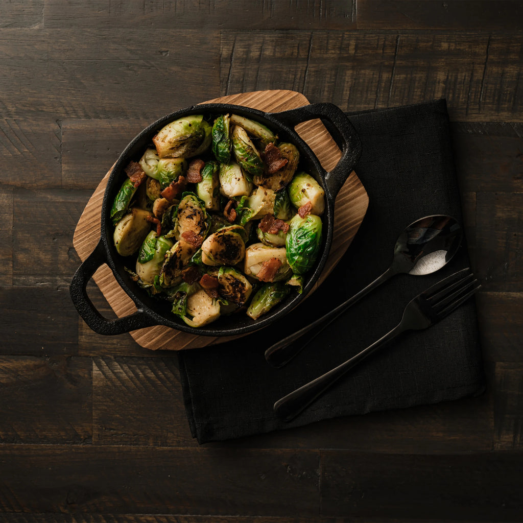 A cast iron bowl full of smoked Brussel sprouts sits on a wooden board which sits next to a spoon and fork on a folded black cloth. All of that sits on a dark-stained wooden table.