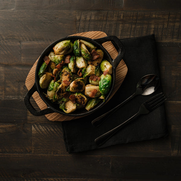 A cast iron bowl full of smoked Brussel sprouts sits on a wooden board which sits next to a spoon and fork on a folded black cloth. All of that sits on a dark-stained wooden table.