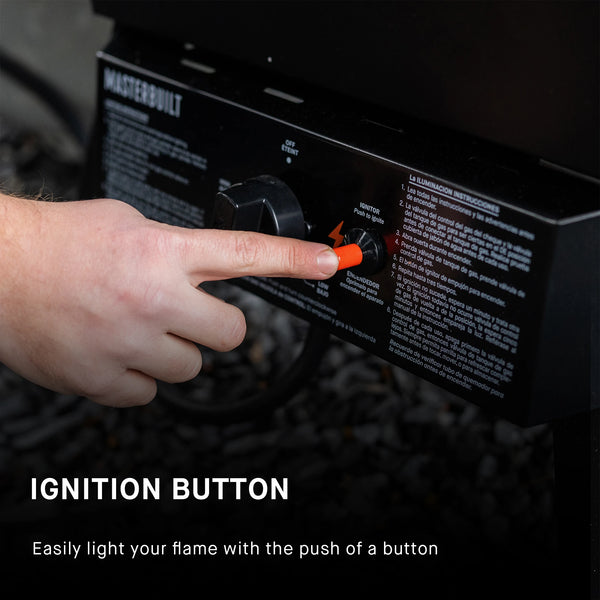 Closeup of a hand pressing an ignition button on a Masterbuilt dual fuel smoker. The button is mounted on the control panel located below the smoker body. The button is to the right of the temperature control dial. Text reads: Ignition button. Easily light your flame with the push of a button.