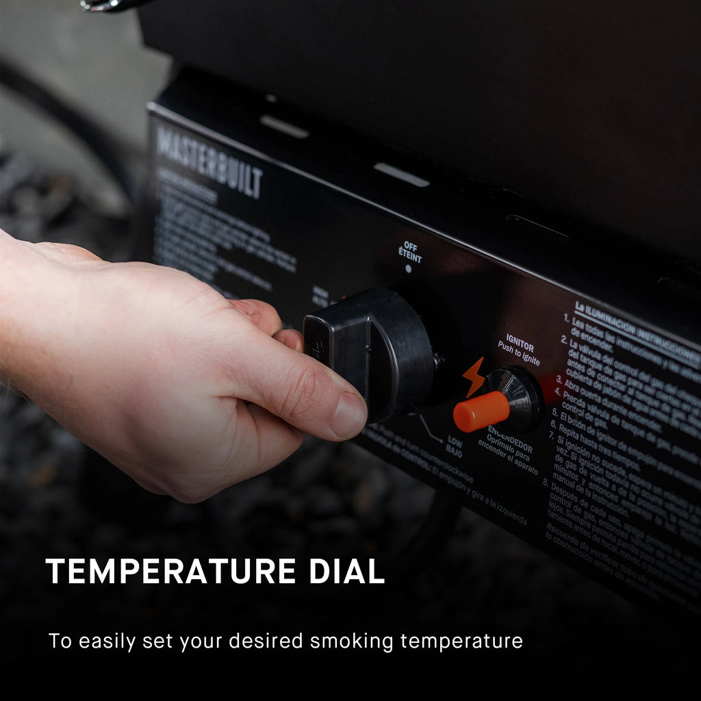 Closeup of a hand adjusting a temperature dial on a Masterbuilt smoker. The dial is mounted in the control panel below the smoker body. Text reads: Temperature dial to easily set your desired cooking temperature.