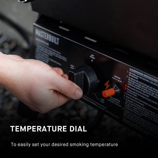 Closeup of a hand adjusting a temperature dial on a Masterbuilt smoker. The dial is mounted in the control panel below the smoker body. Text reads: Temperature dial to easily set your desired cooking temperature.