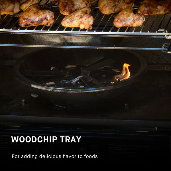 A block of smoking wood buns in the flame disk bowl placed on the bottom of the smoker body. Chicken wings smoke on the rack above it. The removable water bowl hangs above the flame disk bowl and below the smoking rack. Text reads: Woodchip tray for adding delicious flavor to foods.