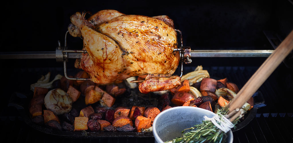A whole chicken roasts on Gravity Series Rotisserie with a pan of vegetables roasting while catching the drippings below. A basting stick with fresh herbs tied to it rests in a bowl of broth on the front shelf of the grill.