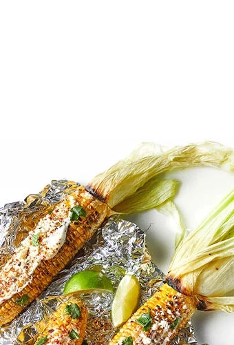 Grilled corn on the cob, with the husks pulled back to make handles, topped with fresh herbs, sour cream, and spices. The corn sits on a wrinkled piece of aluminum foil along with lime wedges.
