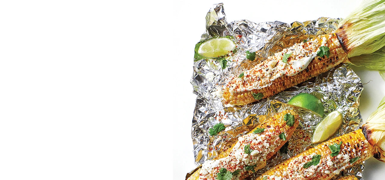 Grilled corn on the cob, with the husks pulled back to make handles, topped with fresh herbs, sour cream, and spices. The corn sits on a wrinkled piece of aluminum foil along with lime wedges.