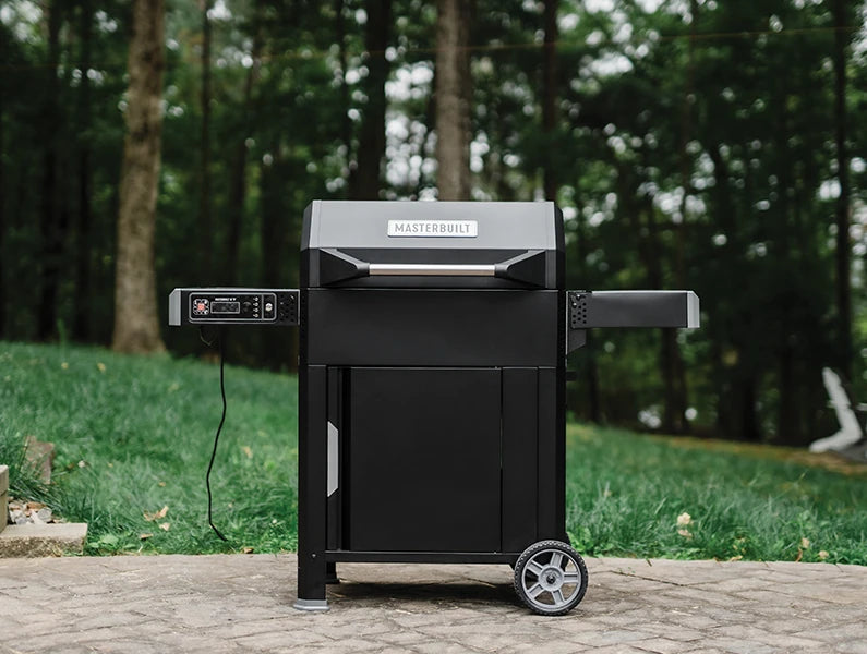 An AutoIgnite Series 545 grill sitting outside on a paved patio in front of a lush green lawn with blurry trees in the background. The grill has an enclosed, wheeled cart and 2 side shelves. The control panel is mounted under the left shelf and has a cord dangling from it.