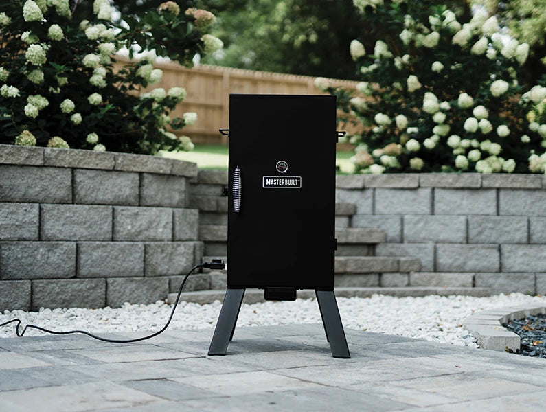 Masterbuilt Electric Smoker with legs sitting on a patio paved with grey blocks. A light grey retaining wall and bushes with clumps of white flowers are behind it.