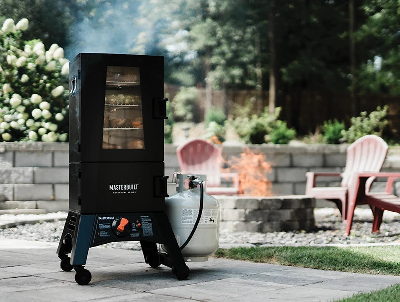 Smoking on the patio. 4 racks of food are visible through the window in the top door. Smoke rises from the top of the smoker. A small propane tank is attached on the right. A handle used to move the smoker is attached to the top left panel of the smoker body. 