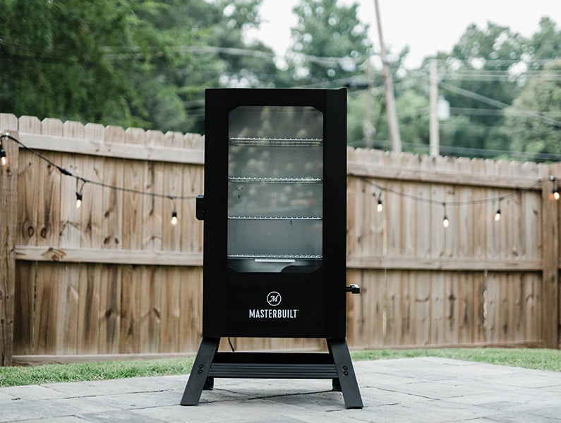 Black Masterbuilt smoker on a patio with a wooden fence and trees in the background. The smoker has a window in the door. The smoker body is filled with smoke.