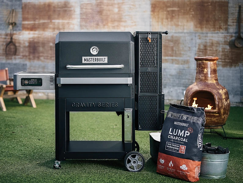 A Gravity Series 1050 grill stands with a bag of Masterbuilt lump charcoal next to it. The bag of charcoal leans against a metal bucket full of charcoal lumps. 